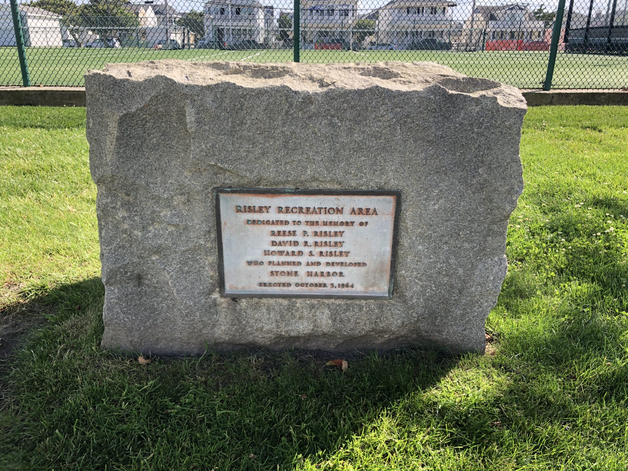 Color photo taken June 2024 showing the same granite monument and bronze ‘Risley Recreation Area’ plaque in bright sunlight on a trimmed lawn, with fenced public tennis courts and shore homes in the background.