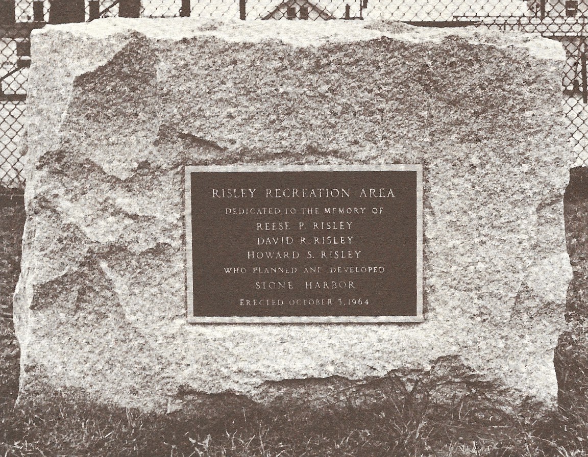 Archival black-and-white image of a rough-hewn granite block bearing a bronze plaque that reads ‘RISLEY RECREATION AREA … Erected October 3, 1964,’ honoring Reese, David and Howard Risley; chain-link tennis-court fence visible behind.