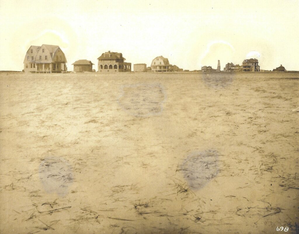 Distant view across open sandy land of Stone Harbor, NJ showing several buildings along a street, including a large gambrel-roofed cottage on the far left, a Mediterranean-style stucco house with a garage, and additional homes and larger buildings receding into the distance.