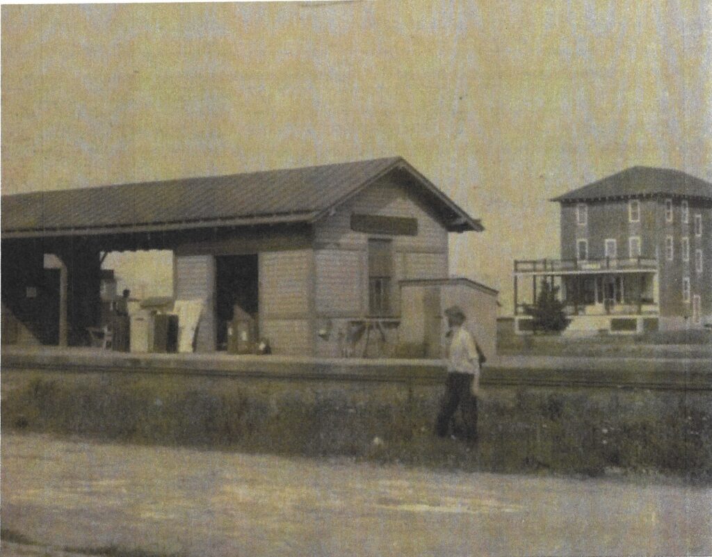 Small railroad station building in the foreground with a man standing nearby, and the Fairview Hotel visible in the background, showing their close proximity in early Stone Harbor.