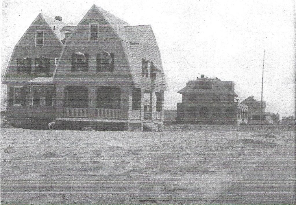  Completed three-story shingled cottage with gambrel roof, porch railings and spindles, shuttered windows with awnings, and a smaller building visible to the right. The image is somewhat faded and low in contrast.