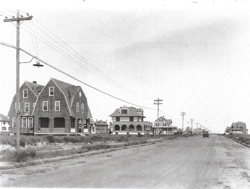 Black-and-white photograph looking north along a wide graveled avenue lined with electric poles and wires. A large completed shingled cottage stands on the left, with additional homes and a stucco building visible along the street and an automobile in the distance.