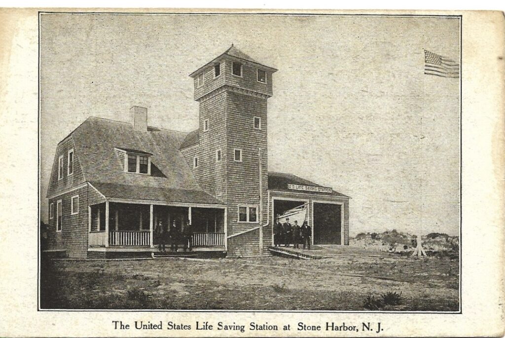 Vintage postcard of the United States Life Saving Station at Stone Harbor, a shingled building with a tall observation tower, covered porch, and large open bay. Several uniformed men stand near the bay, and an American flag is visible in the upper right.