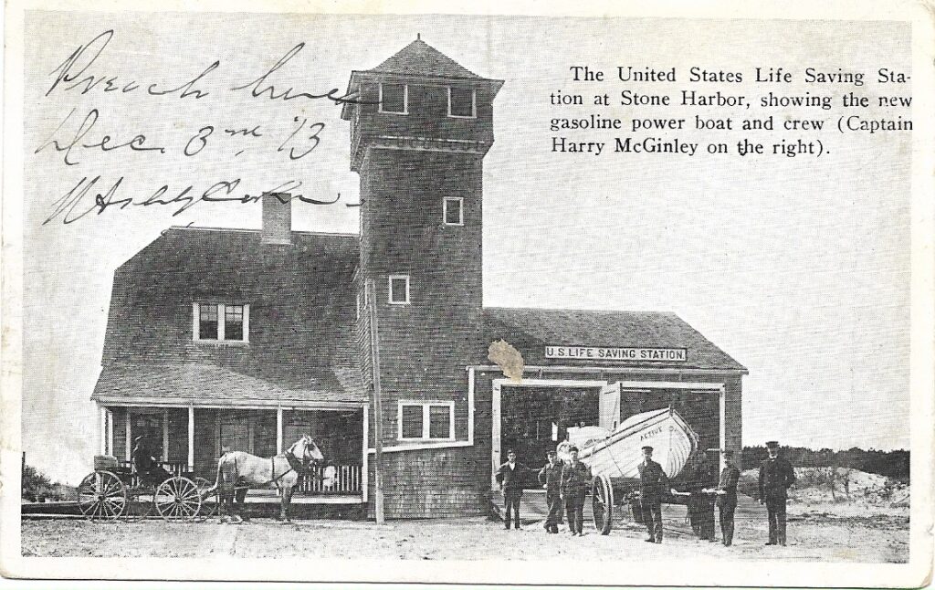 Vintage postcard of the U.S. Life Saving Station at Stone Harbor showing the building with observation tower, a horse-drawn wagon at left, and a surfboat on a wheeled cart at right with several crew members standing nearby. Handwritten inscription and a printed caption identifying Captain Harry McGinley are visible.