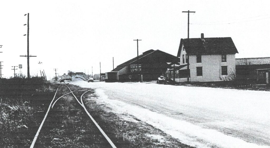 Black-and-white photograph looking east along railroad tracks that split into a spur on the right, with a large lumberyard building and sign visible in the center, a house to the right, utility poles throughout, and a bridge in the distance. Two automobiles are visible on the road.