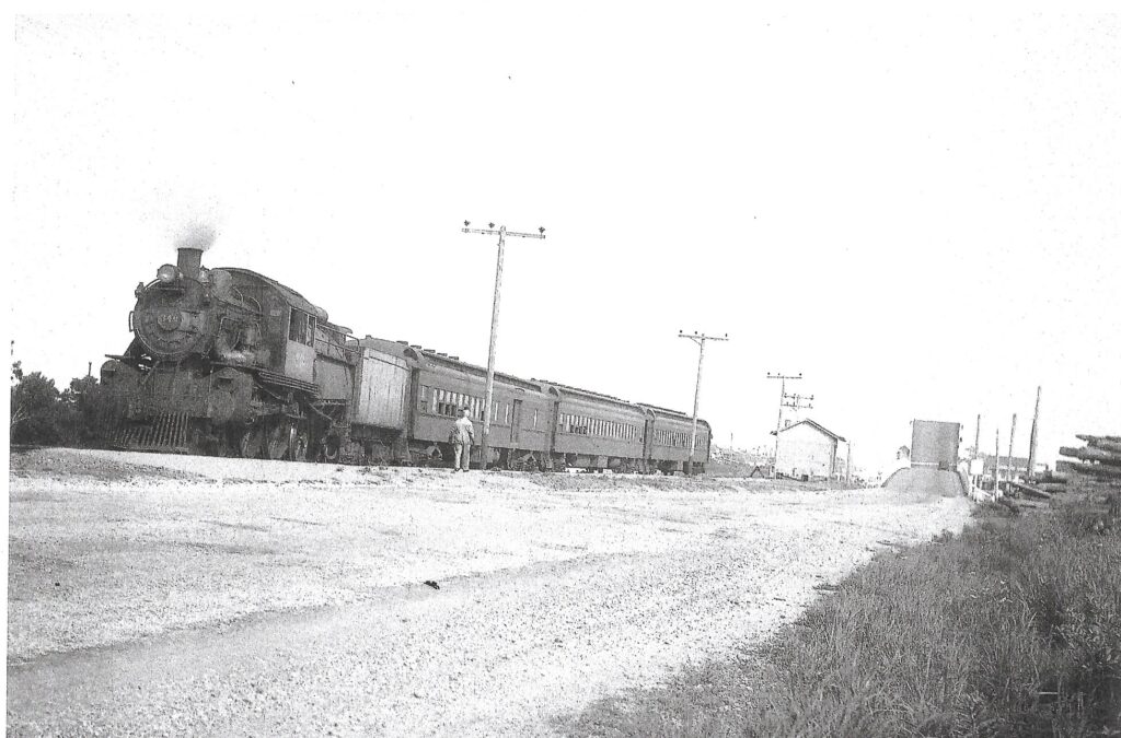 Black-and-white photograph of a steam locomotive and three passenger cars stopped at the end of a rail line, with a person standing near the train, utility poles along the roadway, and a drawbridge visible in the background.