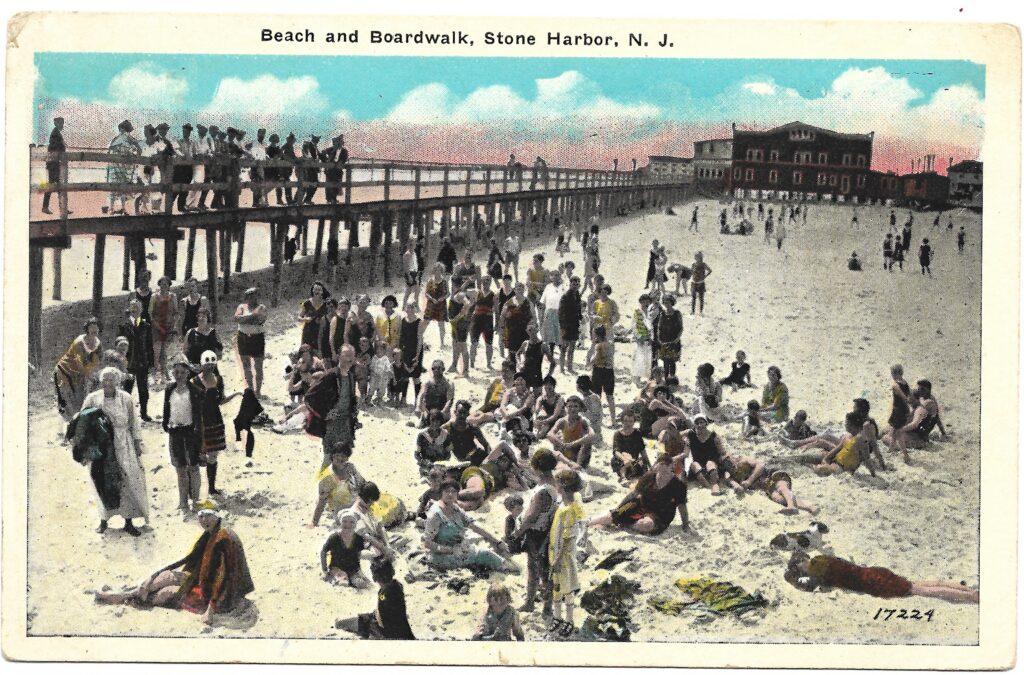 A colorized vintage postcard titled “Beach and Boardwalk, Stone Harbor, N.J.” depicting a large crowd of beachgoers in early 20th-century bathing suits on the sand, with the wooden boardwalk and commercial buildings visible in the background.