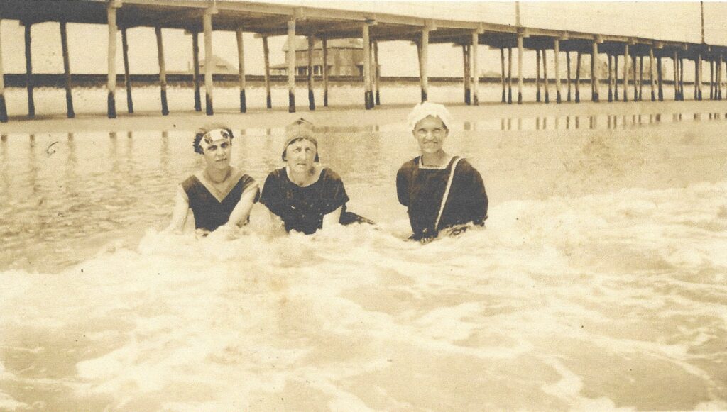A sepia-toned photograph of three women in dark bathing suits and swim caps sitting together in the surf, smiling at the camera, with the wooden pilings of the Stone Harbor boardwalk visible behind them.
